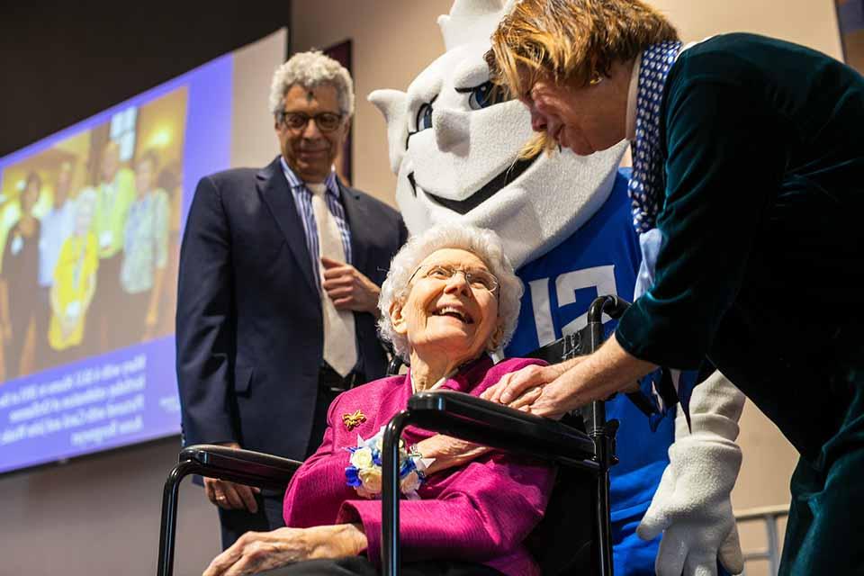 Dr Fran Pestello holds Mary Bruemmer's hand at her 100th birthday celebration while the Billiken and Dr Fred Petesllo look on